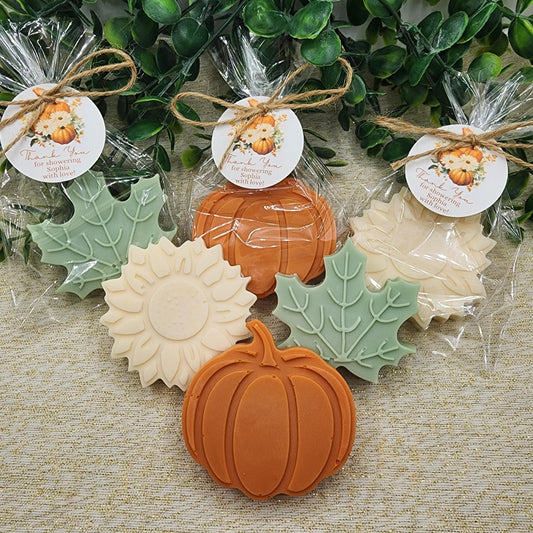 Decorative soaps shaped like pumpkins, leaves, and sunflowers on a textured surface with greenery in the background.