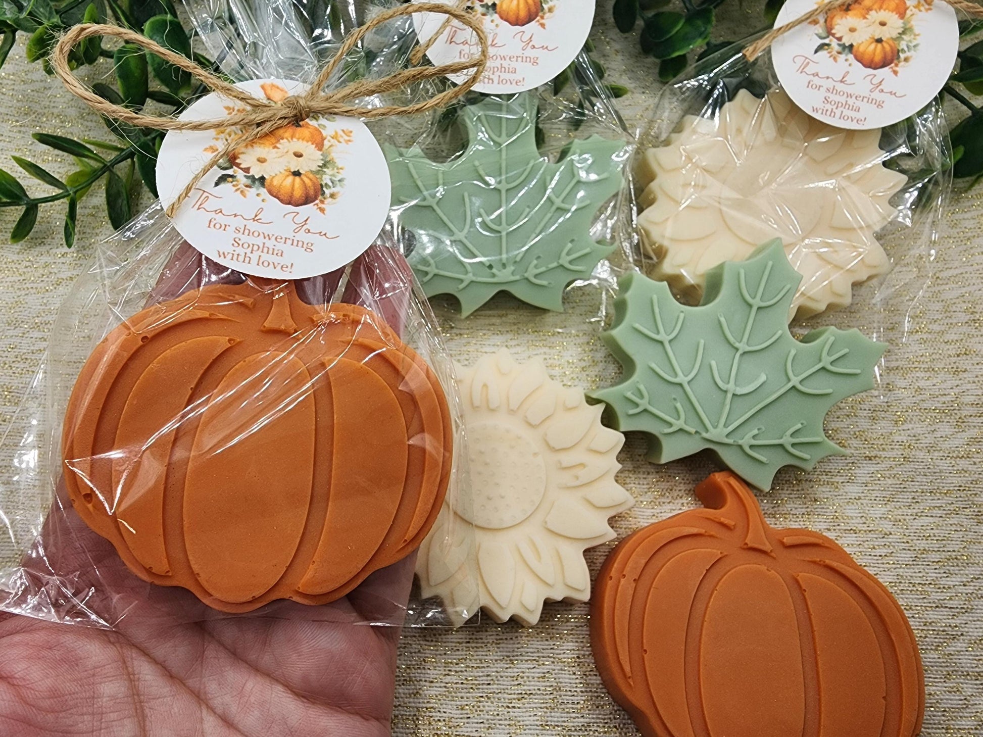 Pumpkin, leaf, and round soap bars in clear packaging with labels on a textured surface.