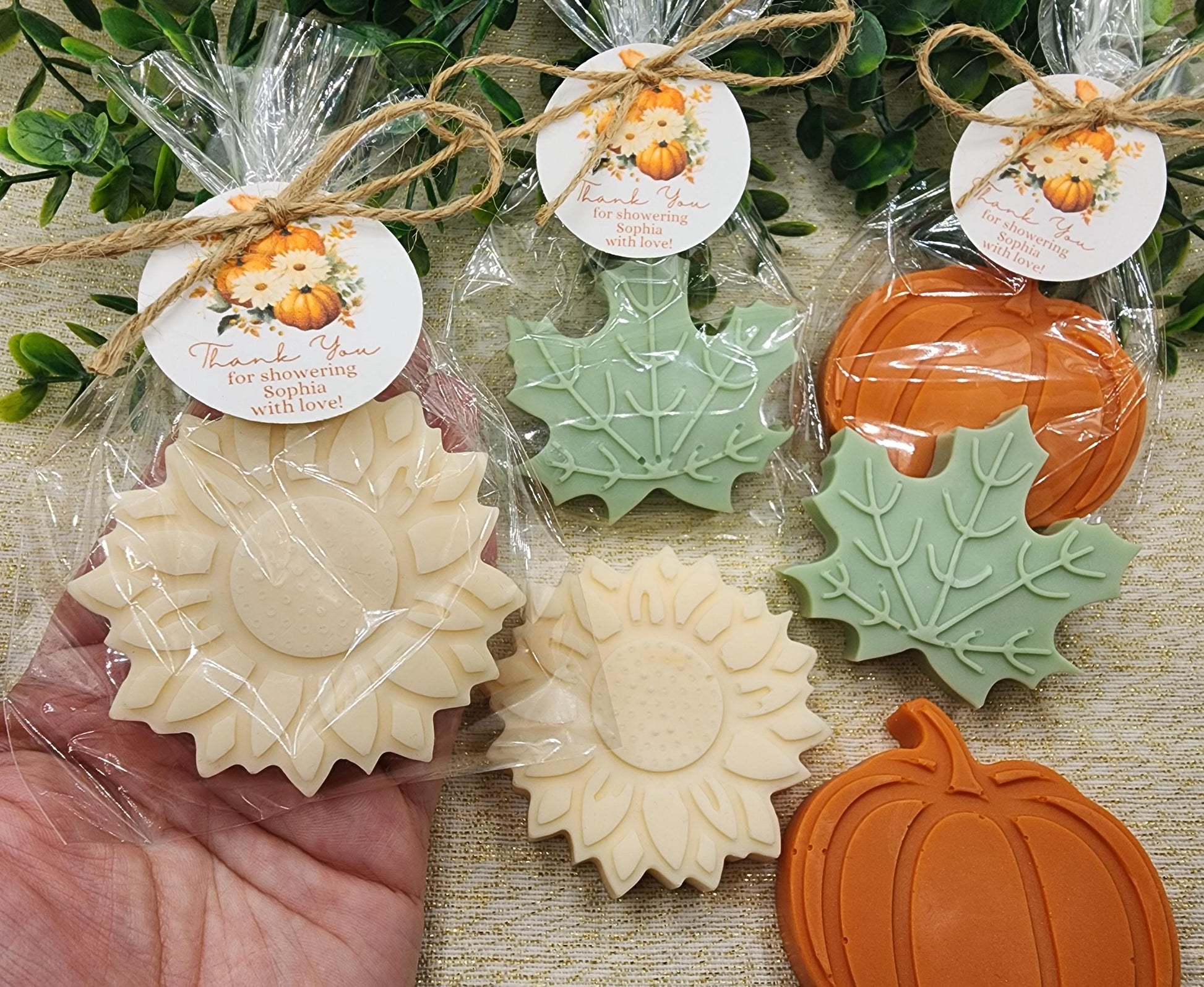 Decorative soap bars shaped like pumpkins and leaves, wrapped in cellophane with tags, on a textured surface.