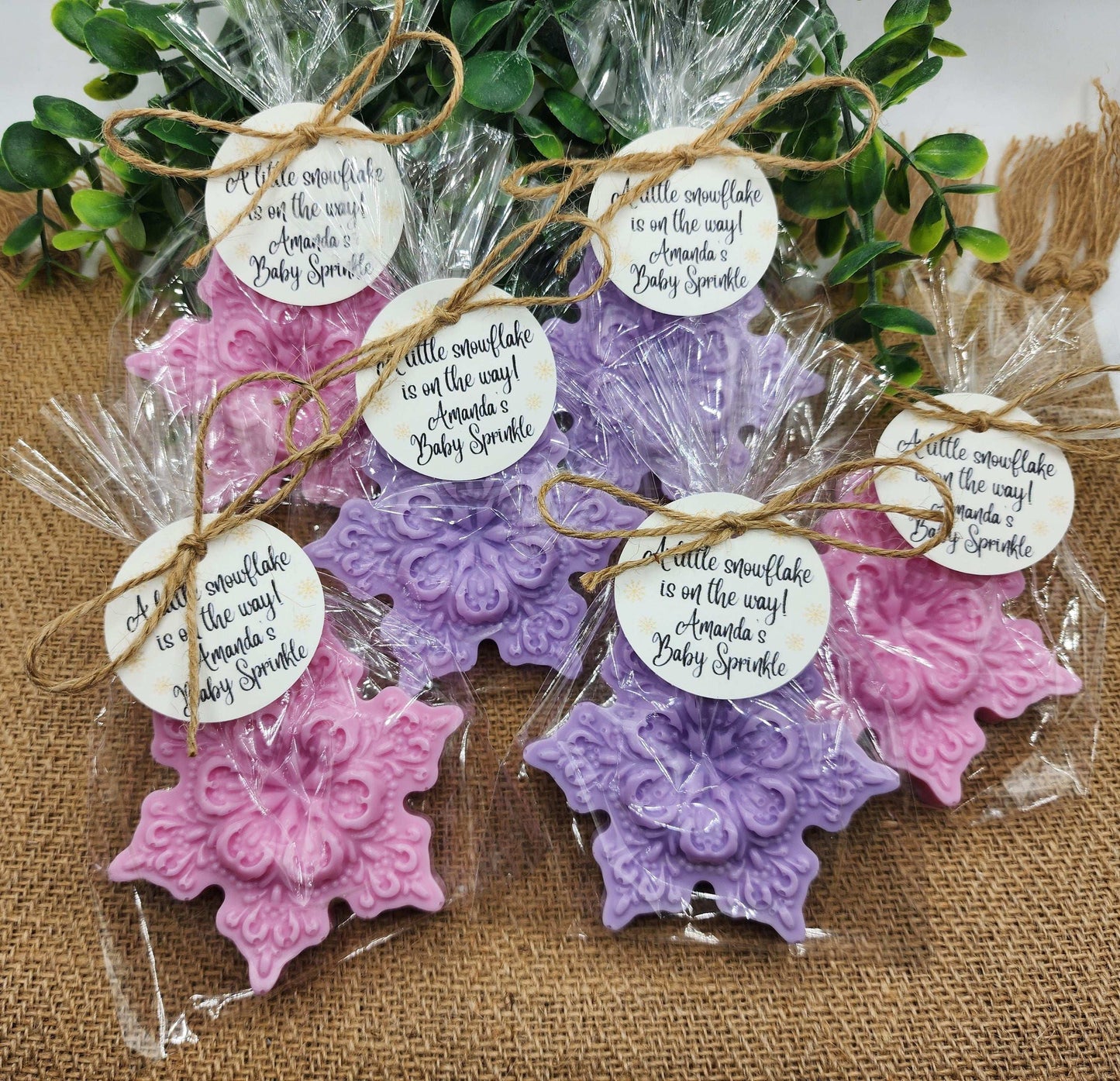 Decorative snowflake-shaped favors in pink and purple with tags on a burlap surface.