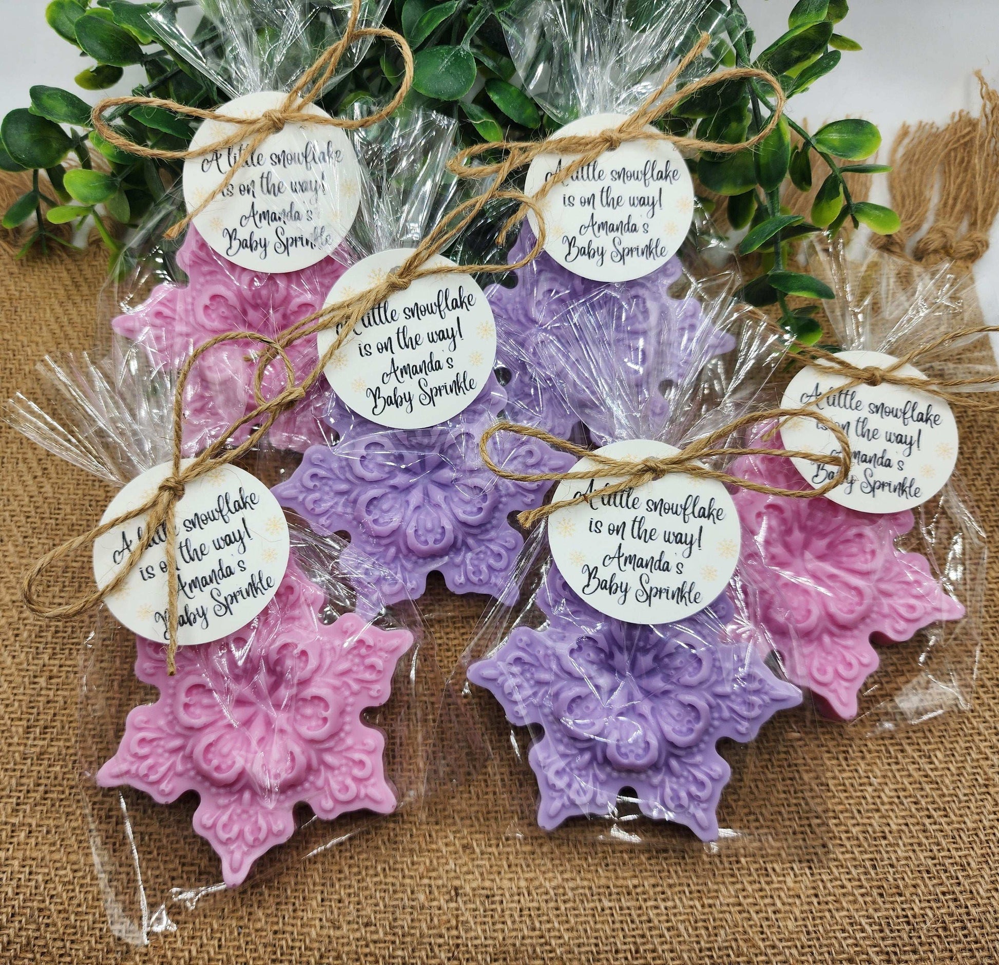 Decorative snowflake-shaped favors in pink and purple with tags on a burlap surface.